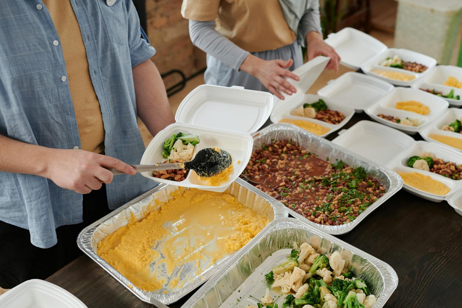 Volunteers plating warm, fresh meals into takeaway containers at a community kitchen, hands and food in focus.