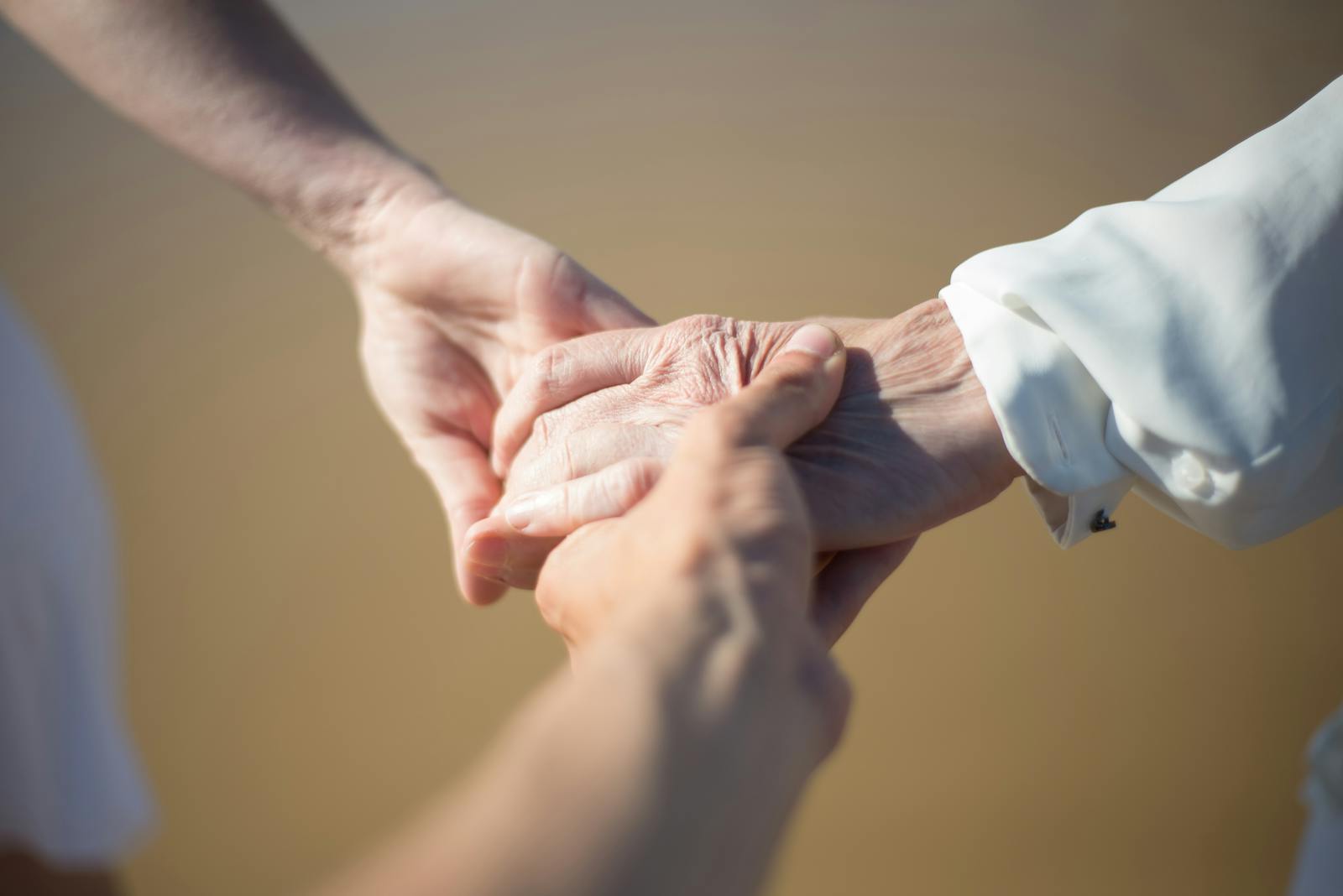 Two pairs of hands clasped gently together against a soft, warm sand-coloured background.