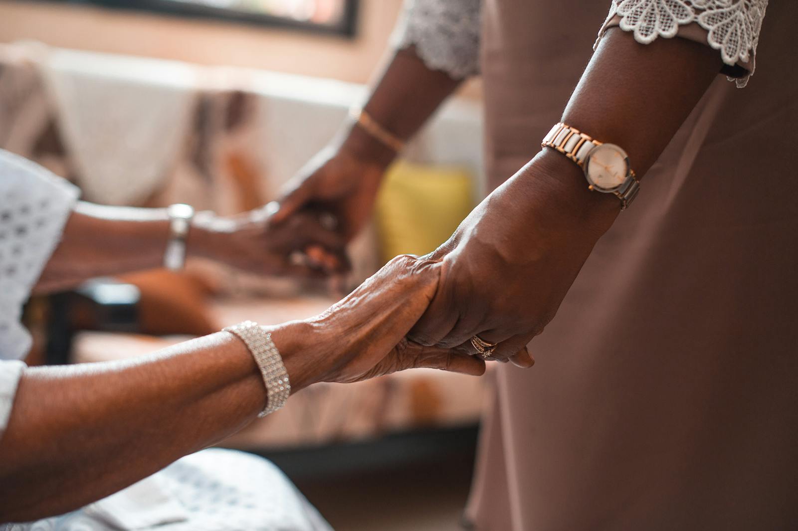 Two people holding hands warmly, close-up, with gold and pearl jewellery and soft indoor light.