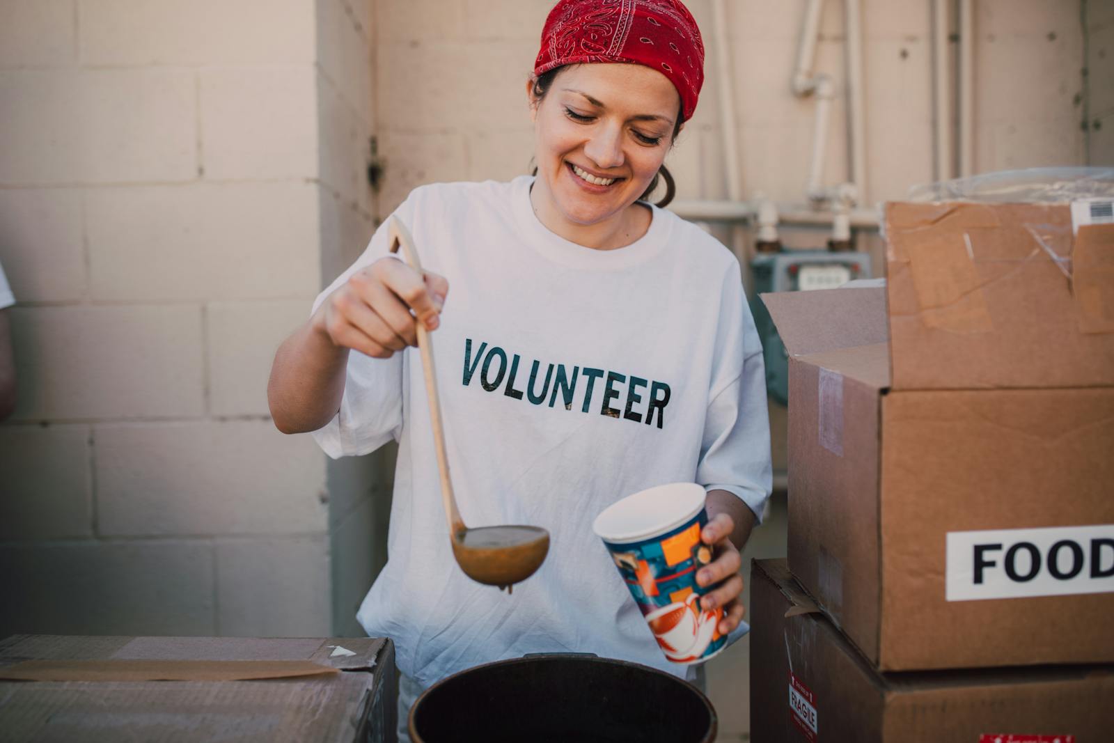 A smiling volunteer in a red headscarf ladles hot soup into a cup beside boxes marked food at a donation point.